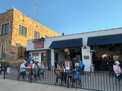 A lively taco restaurant with outdoor seating, people dining, and a bright "Open" sign on a sunny day.