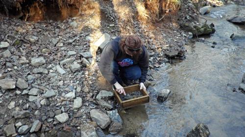 A person crouches by a stream, sifting through rocks in a wooden box, surrounded by a rocky landscape.