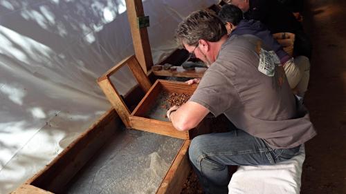 A person sifts through materials in a wooden box at a workbench, focused on their task in a well-lit space.