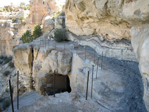 A rocky cliffside with a narrow path leading to a cave entrance, surrounded by natural formations and sparse vegetation.