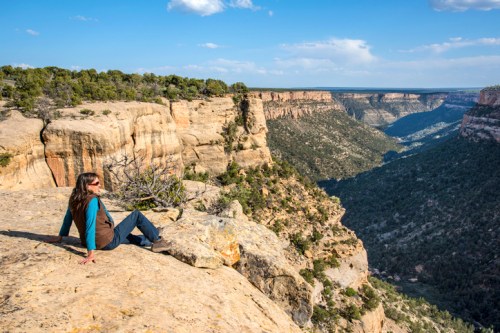 A person sits on a rocky ledge, overlooking a vast canyon landscape under a clear blue sky.