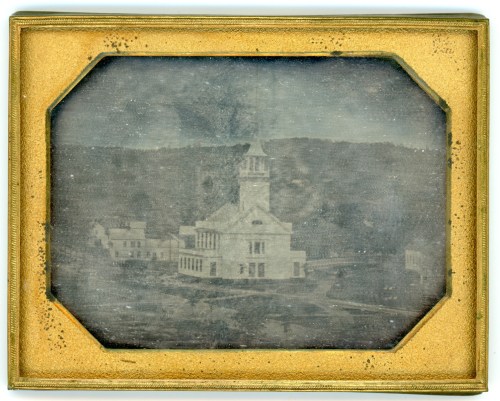 Historic black-and-white image of a building with a clock tower, surrounded by trees and hills in the background.
