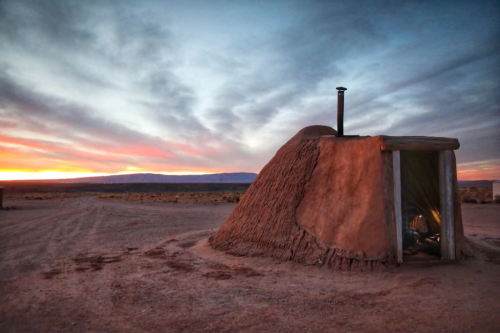 A traditional adobe structure at sunset, with a chimney and a colorful sky in the background.