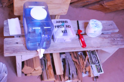 A wooden bench with a water dispenser, first aid kit, lighter, and firewood in a rustic setting.