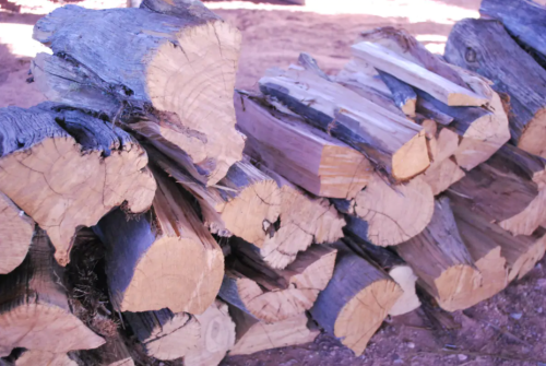 Stacked logs of various sizes, showcasing their natural wood grain and cut ends, resting on a sandy surface.