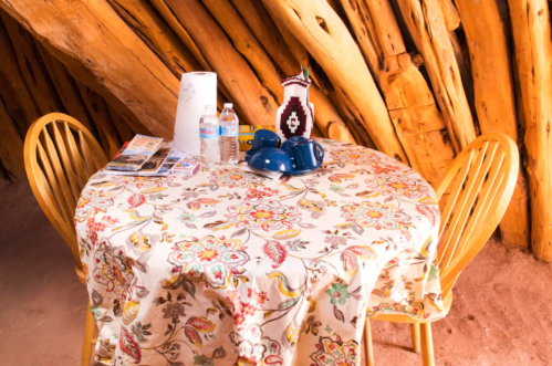 A wooden table with a floral tablecloth, two blue teapots, water bottles, and a decorative vase in a rustic setting.