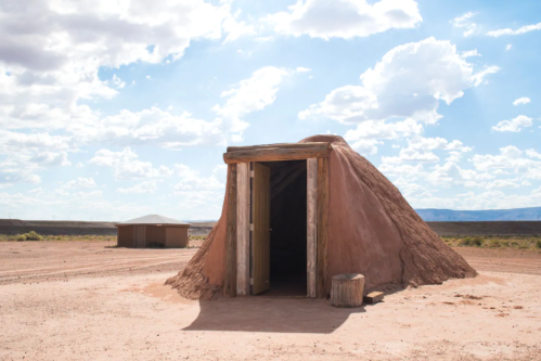 A traditional adobe structure with a wooden door, set against a blue sky with clouds in a desert landscape.