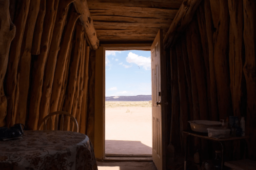 Interior of a rustic wooden hut with an open door, revealing a desert landscape and blue sky outside.