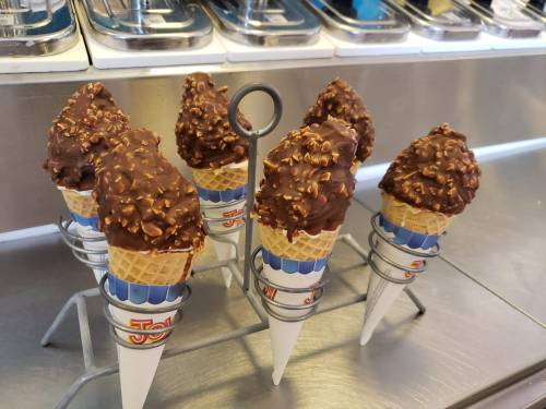 Six chocolate-dipped ice cream cones with crunchy toppings displayed in a metal holder on a counter.