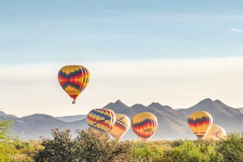 Colorful hot air balloons float over a desert landscape with mountains in the background under a clear blue sky.