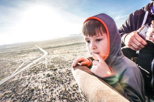 A young boy in a hoodie gazes thoughtfully over the edge of a hot air balloon, with a vast desert landscape below.