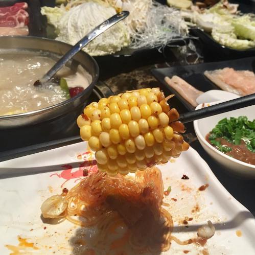 A skewer holds a piece of corn above a plate with noodles and various ingredients, with a hot pot in the background.