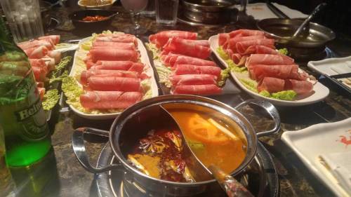 A table set with plates of thinly sliced meat and a hot pot filled with broth, surrounded by condiments and drinks.