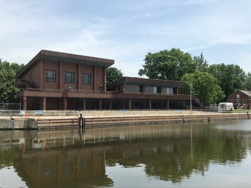 A modern brick building by a calm waterway, surrounded by trees and reflecting in the water.