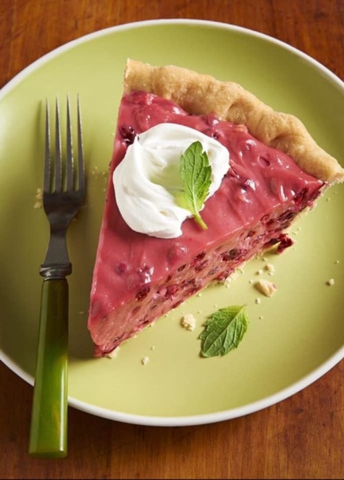 A slice of cherry pie topped with whipped cream and a mint leaf, served on a green plate with a fork.
