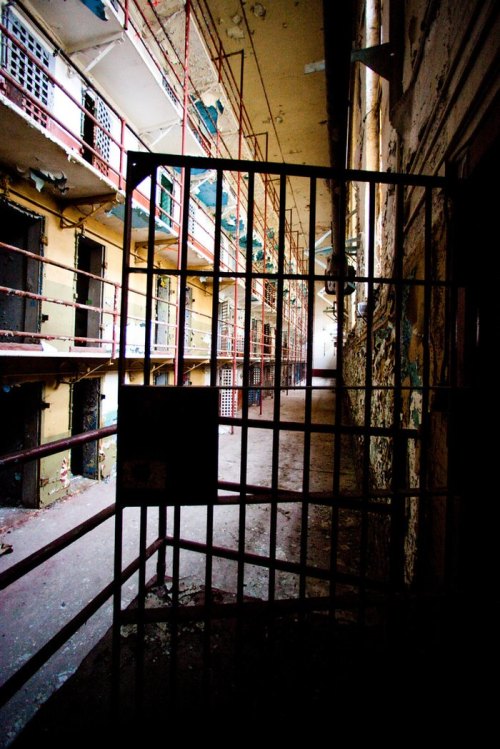 A dimly lit, abandoned prison corridor with peeling walls and barred cells on either side.