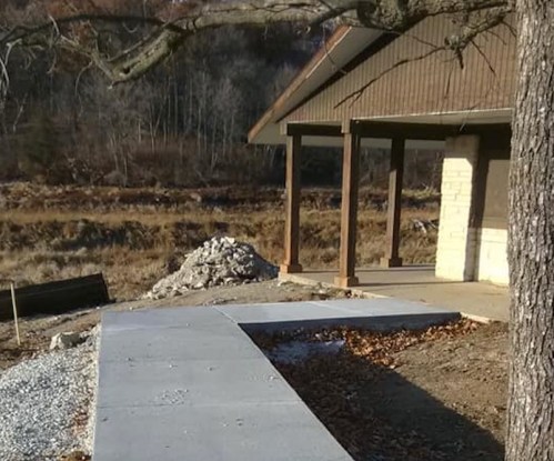 A concrete walkway leads to a house, with a pile of gravel nearby and a wooded area in the background.