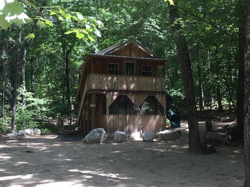 A wooden two-story cabin surrounded by trees and rocks in a forested area.