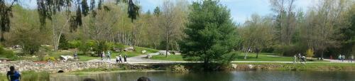 A serene park scene with a pond, walking paths, trees, and people enjoying the outdoors on a sunny day.