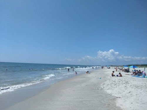 A sunny beach scene with people enjoying the water and sand, under a clear blue sky with a few clouds.