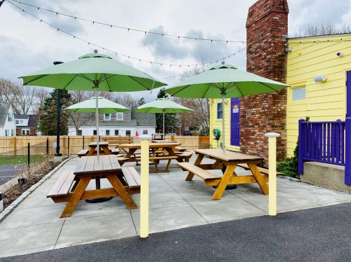 Outdoor seating area with wooden picnic tables and green umbrellas, beside a yellow building and a brick chimney.