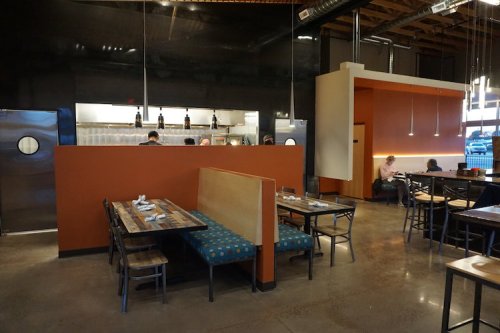 Interior of a modern restaurant featuring wooden tables, a blue booth, and an open kitchen area.
