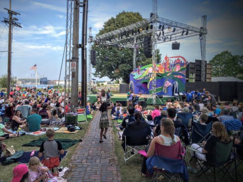 A large crowd enjoys an outdoor performance on a colorful stage, with children playing and people seated on blankets and chairs.