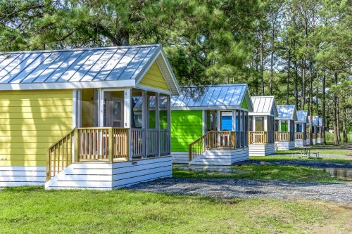 Colorful cabins with porches lined up in a wooded area, surrounded by green grass and tall trees.