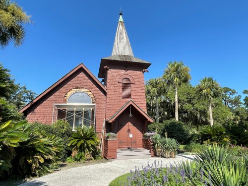 A charming red church with a tall spire, surrounded by lush greenery and palm trees under a clear blue sky.