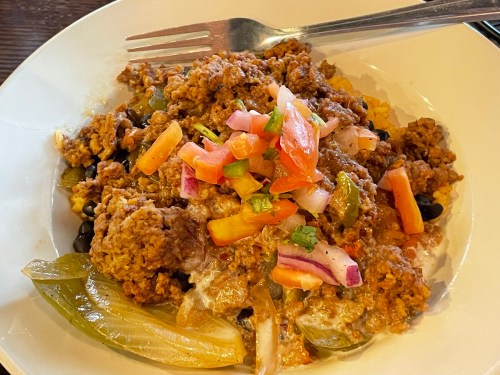 A bowl of seasoned ground meat topped with fresh salsa, served over rice and beans with vegetables.
