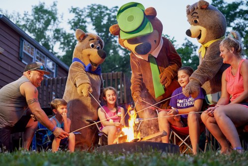 A group of people and two bear mascots gather around a campfire, roasting marshmallows and enjoying the evening.