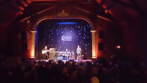 A live music performance on stage with musicians playing instruments in a warmly lit venue filled with an audience.