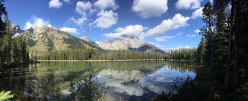 A serene lake surrounded by mountains and trees, reflecting the blue sky and fluffy clouds above.