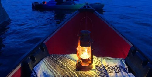 A lantern glows in a kayak, with a person paddling in the background against a twilight sky and rocky shoreline.
