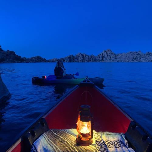A lantern glows in a kayak, with a person paddling in the background against a twilight blue sky and rocky shoreline.