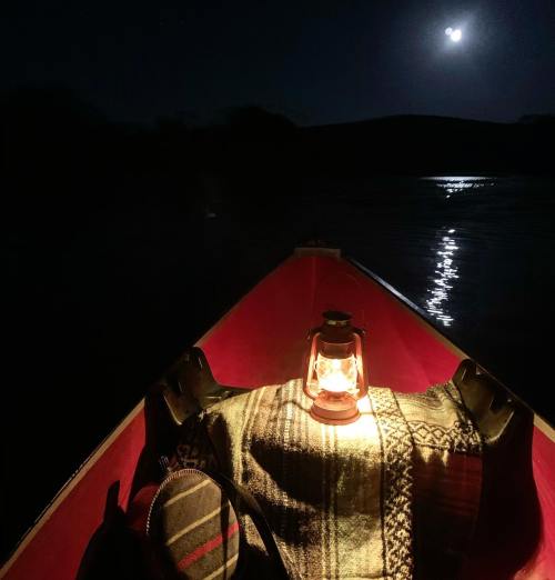 A canoe at night with a glowing lantern on a blanket, reflecting moonlight on the water.