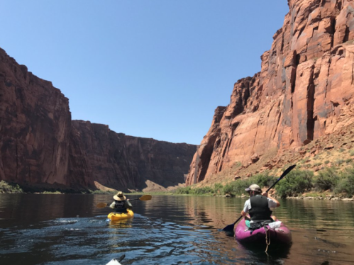Two kayakers paddle through a narrow canyon with towering red rock cliffs under a clear blue sky.
