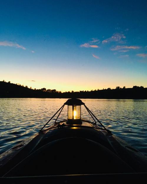 A lantern on a boat illuminates the water at sunset, with silhouettes of trees and hills in the background.