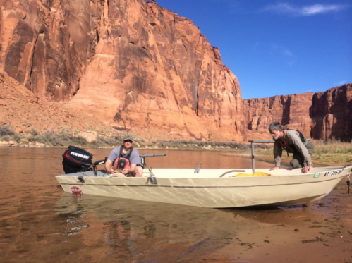 Two men in a small boat on a river, surrounded by towering red rock cliffs under a clear blue sky.