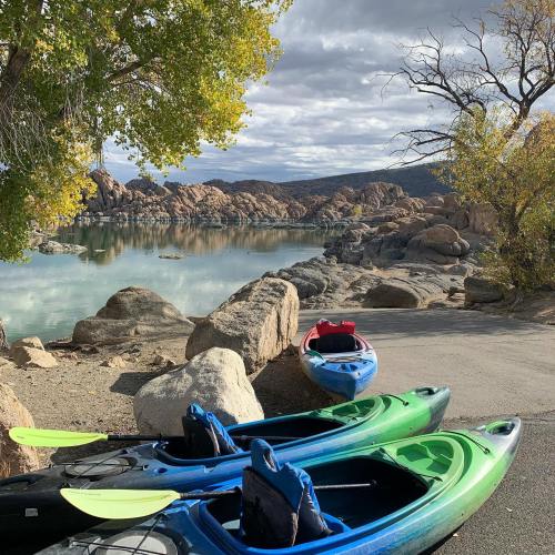 Colorful kayaks lined up by a serene lake, surrounded by rocky shores and autumn foliage under a cloudy sky.