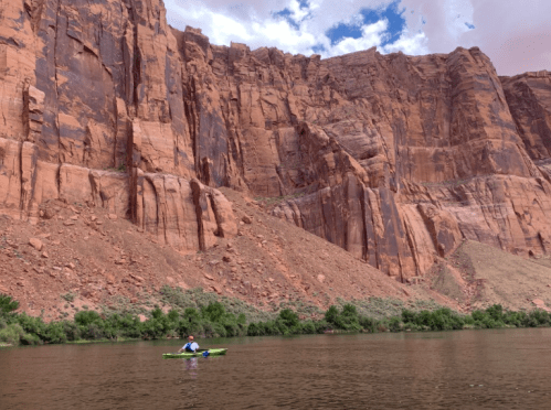 A kayaker paddles on a calm river, surrounded by towering red rock cliffs and lush greenery.