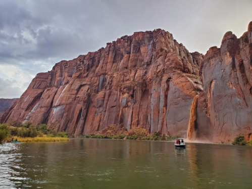 A boat on a river near towering red rock cliffs under a cloudy sky. Water cascades down the rocks in the background.