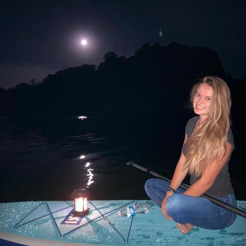 A young woman sits on a paddleboard at night, smiling under a full moon, with a lantern and water nearby.