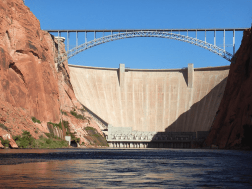 A large dam with a curved bridge above, surrounded by red rock formations and a river in the foreground.