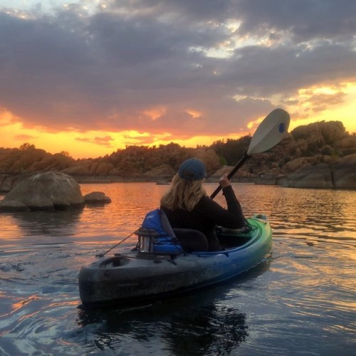 A person kayaking on a calm lake at sunset, surrounded by rocky formations and colorful clouds.