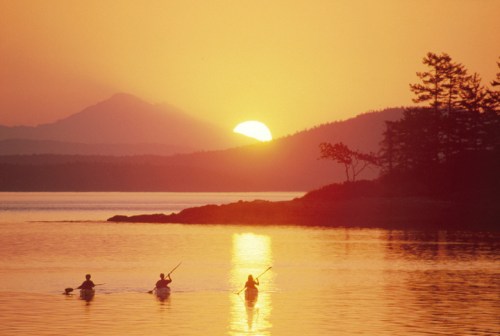 Three kayakers paddle across a calm lake at sunset, with mountains and trees silhouetted against the golden sky.