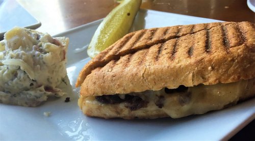 A grilled sandwich with melted cheese and meat, served with a side of coleslaw and a pickle slice on a white plate.