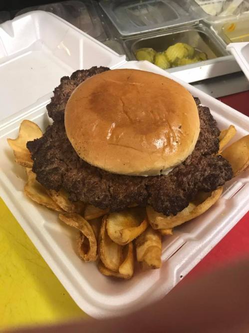 A large hamburger patty on a bun, served on a bed of golden, crispy fries in a takeout container.