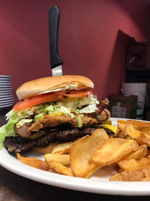 A towering burger with lettuce, tomato, and pickles, served with crispy potato chips on the side.
