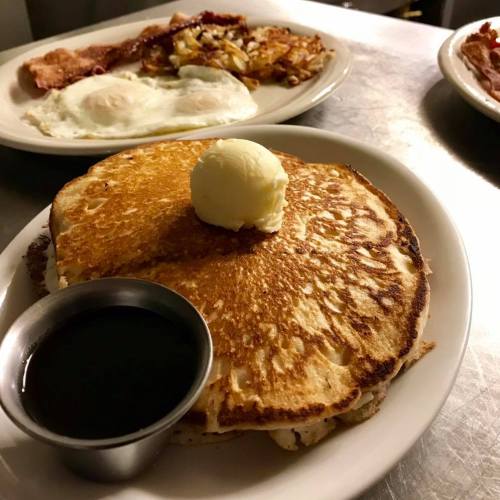 A golden pancake topped with a pat of butter and served with a small cup of syrup, with eggs and bacon in the background.